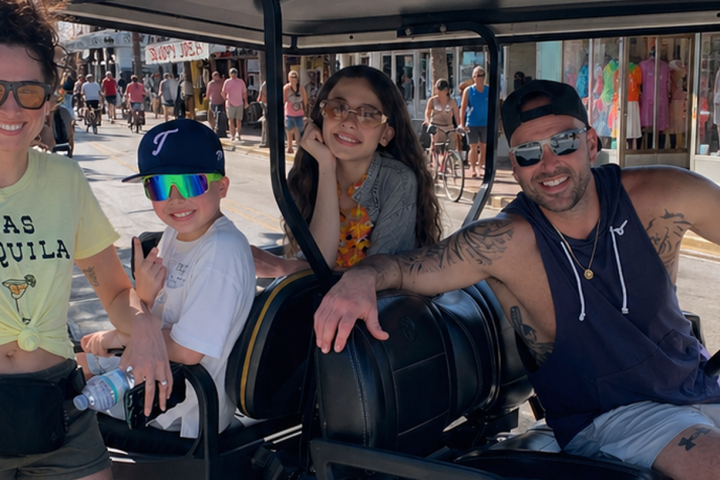 Four people smiling on a golf cart in a sunny street scene.