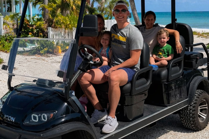 Family on a golf cart by a beach on a sunny day, with palm trees in the background.