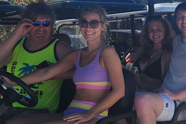 Four people sitting on a golf cart, smiling, in casual summer attire.