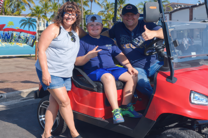 Three people posing with a red golf cart on a sunny street.