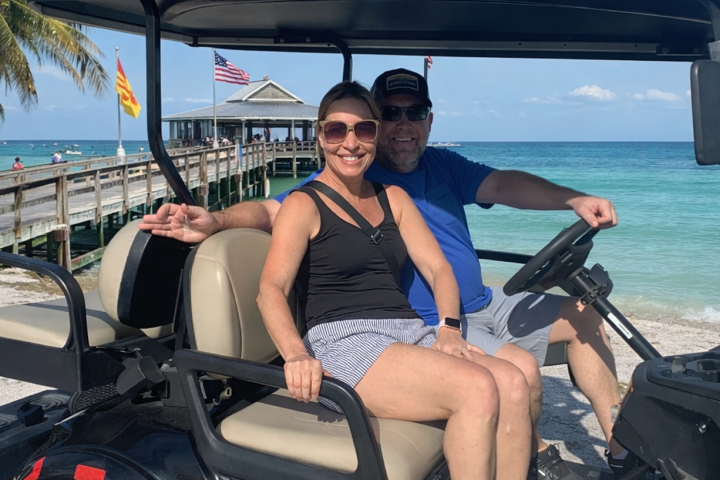 Two people smiling on a golf cart by a beach and pier.