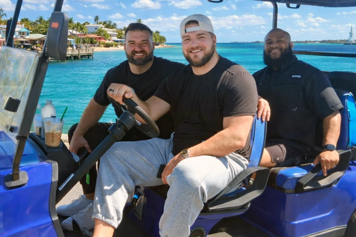 Three men smiling in a blue golf cart by the ocean.