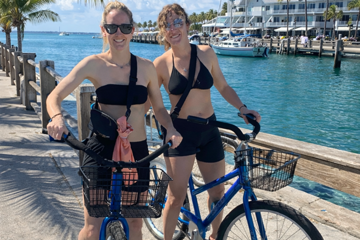 Two women in swimsuits with bikes by the ocean on a sunny day.