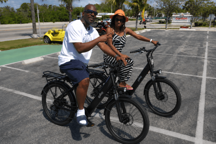 Two people smiling and sitting on black electric bikes in a parking lot.